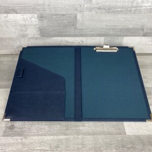 Inside of a navy clipboard folder displayed open to see the crocodile clip on the inside back cover, the shaped pocket and half pocket with a pen strap on the inside front cover and the matching book cloth lining
